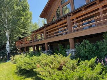 Wooden cabin with a large deck surrounded by greenery on a sunny day.