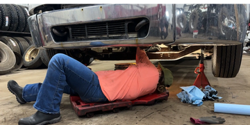 Person in orange shirt working under a lifted vehicle in a garage.