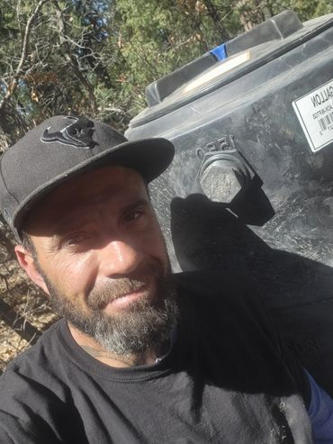 Man with a beard wearing a black cap and shirt outdoors.