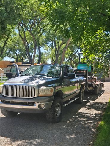A green pickup truck with a trailer parked under trees on a sunny day.
