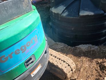 Green excavator with 'Gregory the plumber' near a large black tank outdoors.