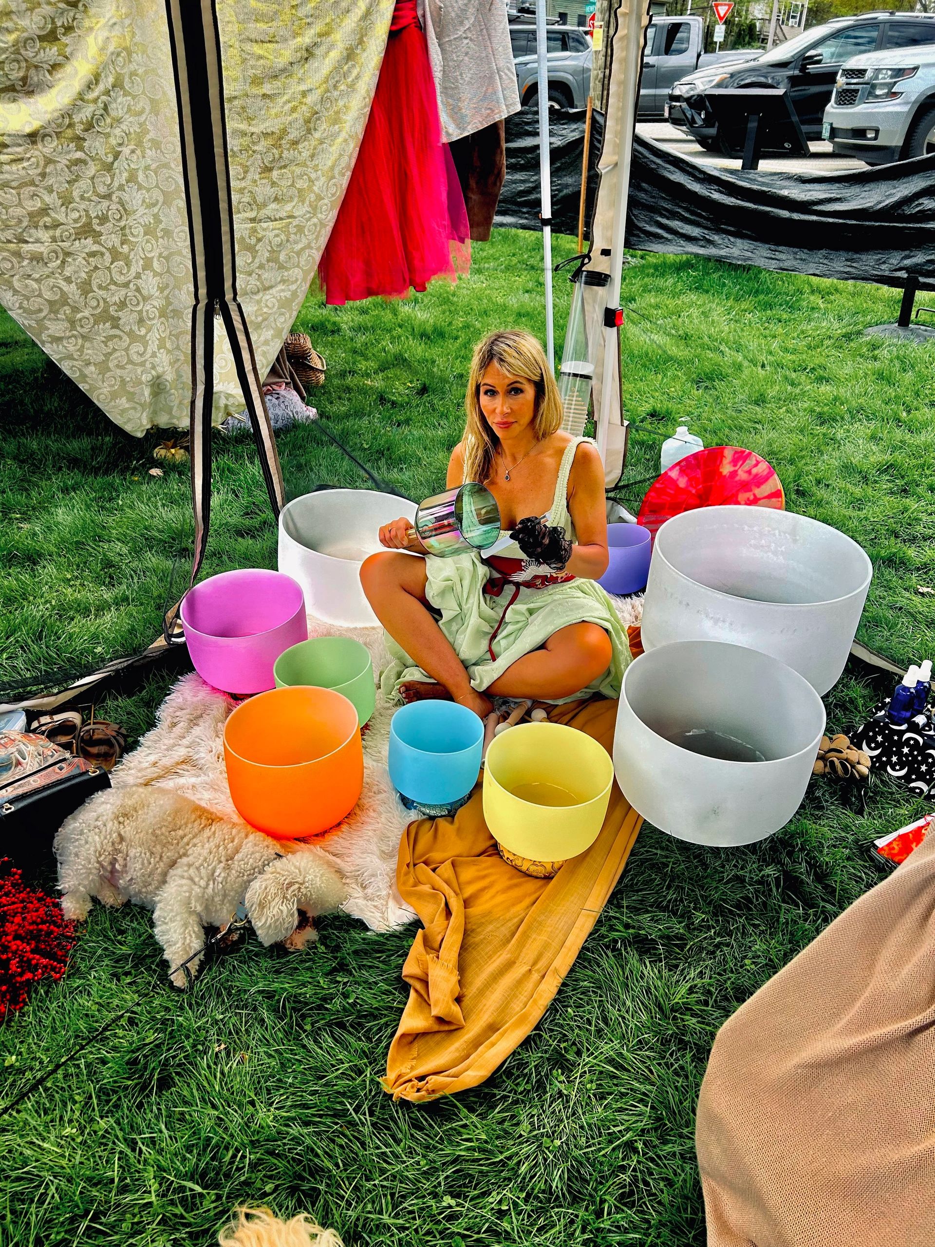 Woman sitting with colorful singing bowls and a small dog on grass.