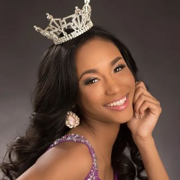 A smiling woman wearing a jeweled crown and purple dress.