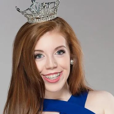 Young woman with red hair wearing a silver crown and blue dress smiling.