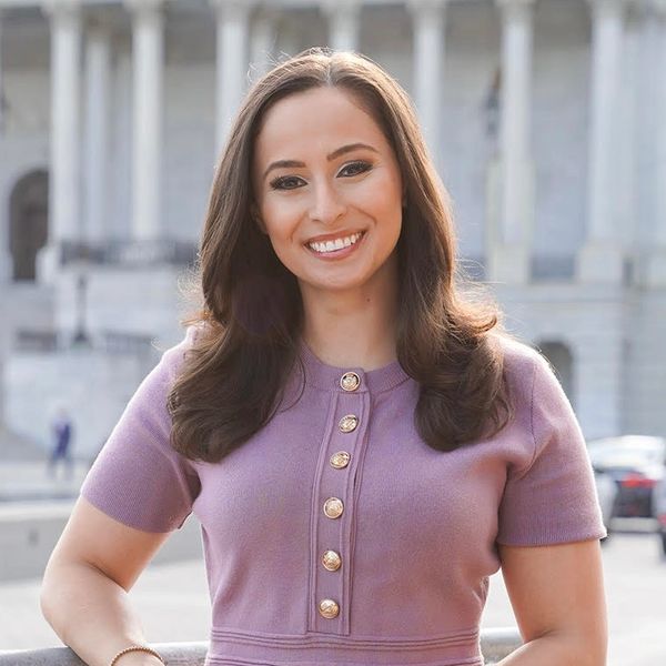 Smiling woman in a purple dress standing outdoors with a historic building in the background.