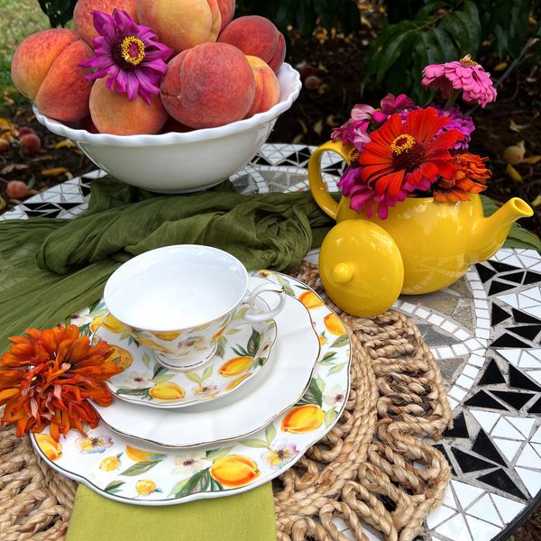A vibrant outdoor tea setting with peaches, floral china, and a yellow teapot filled with flowers.