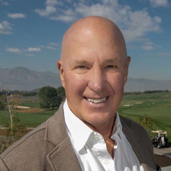 Smiling bald man in a suit outdoors with a golf course background.