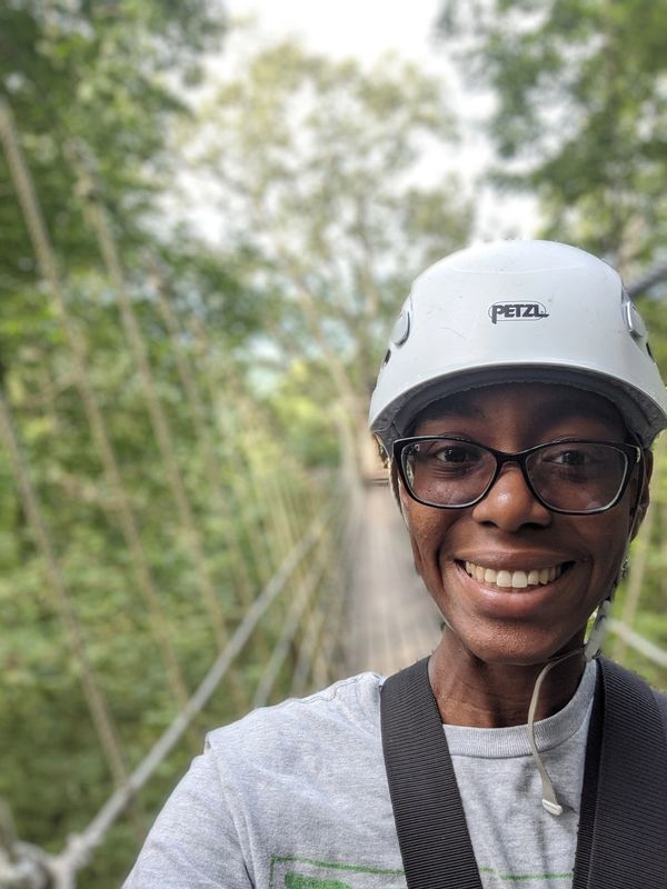 Smiling person wearing a helmet on a suspension bridge in a forest.