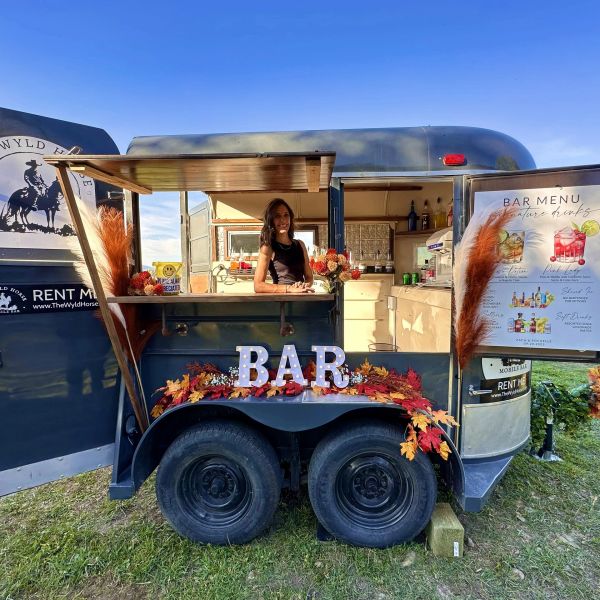 A woman serving drinks from a rustic mobile bar decorated with autumn leaves and a glowing 'BAR' sign.