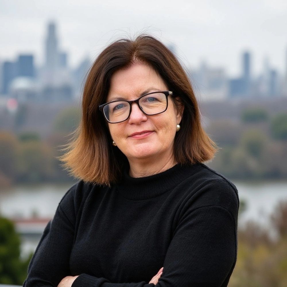 A woman in a black jumper is wearing glasses and smiling, with an urban skyline in the background.