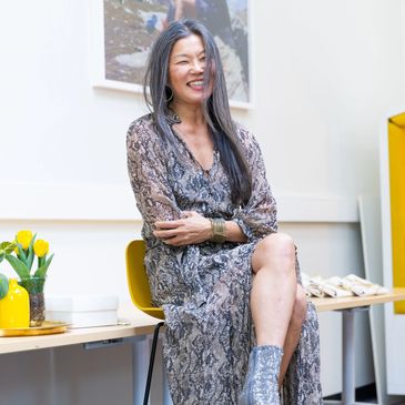 Smiling woman in a patterned dress sitting on a yellow chair indoors.