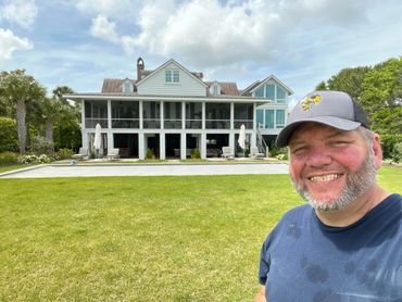 Man smiling in front of a large, white house with a green lawn.