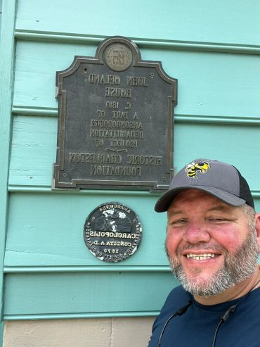 Smiling man in a cap posing by historical plaques on a teal wooden wall.