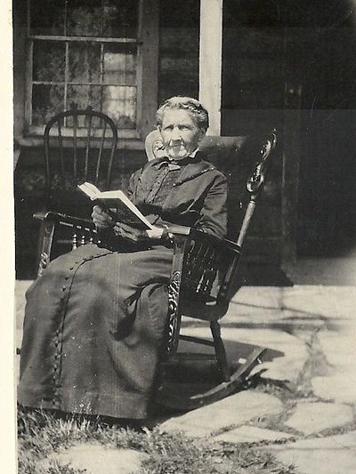Elderly woman reading in a rocking chair outside a house.
