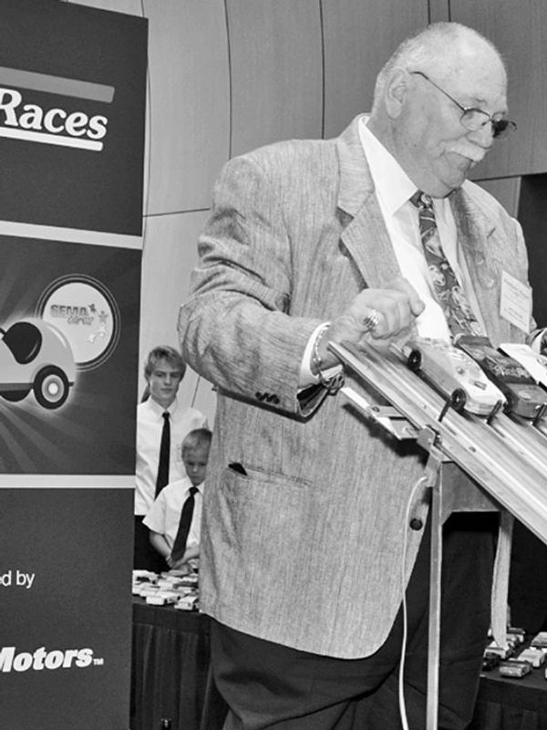 An elderly man in a suit overseeing a model car race event indoors.
