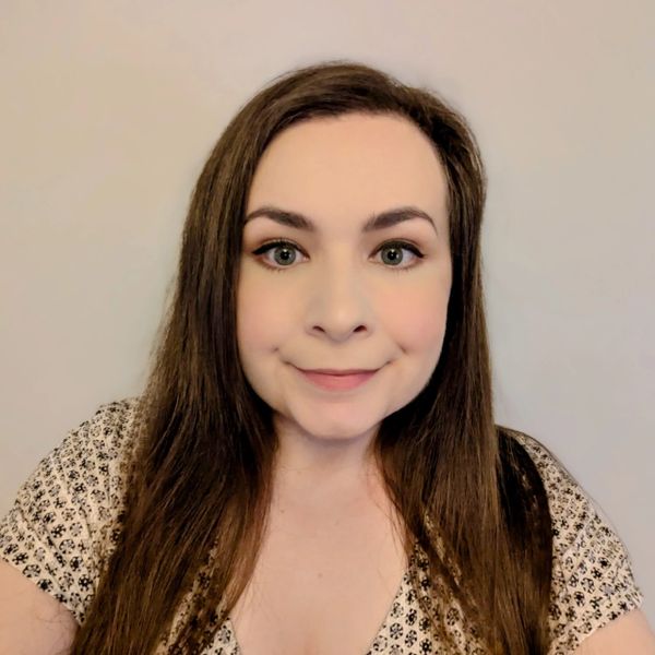 A woman with long brown hair smiling softly against a plain background.