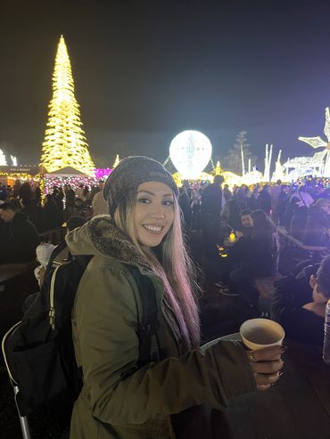 Woman smiling and standing holding hot chocolate amongst a crowd with a tall lit Christmas tree.