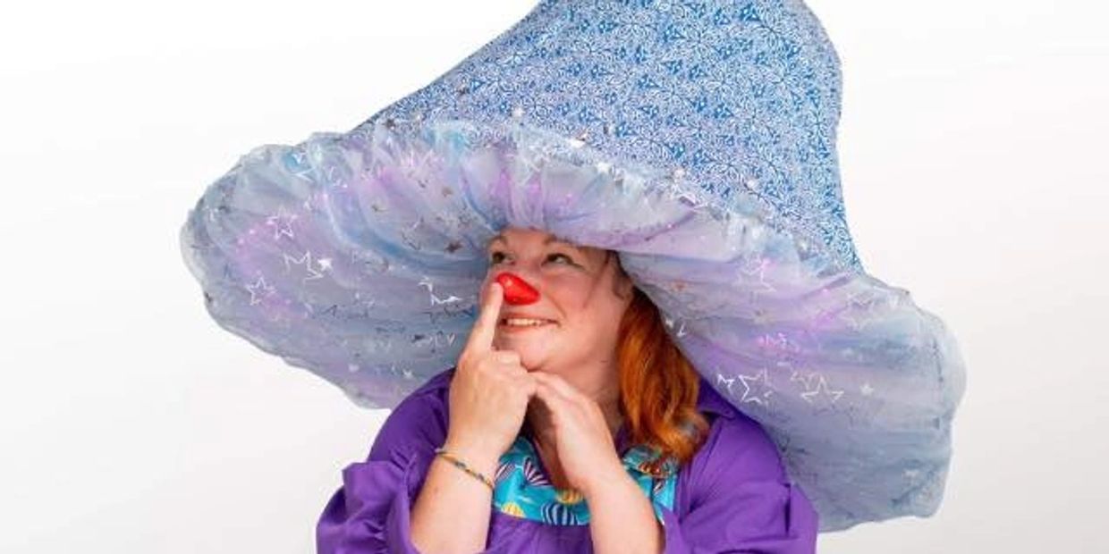 Person wearing a large whimsical blue hat and a red clown nose, smiling playfully.