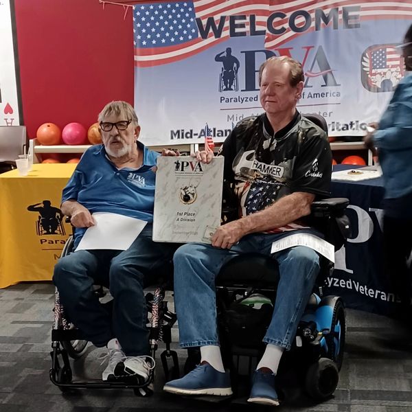 Two men in wheelchairs holding a 1st place certificate at a PVA event.