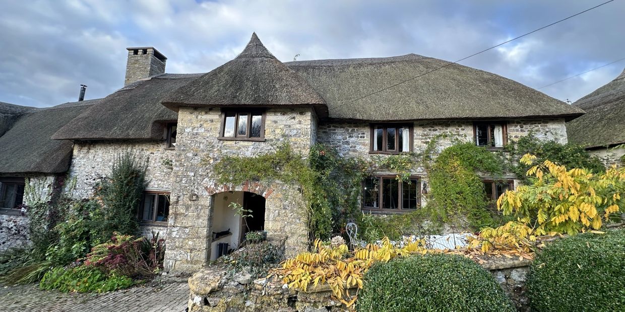 Charming stone cottage with thatched roof and lush greenery under a cloudy sky.
