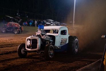 A modified vintage Ford truck racing on a dirt track at night.