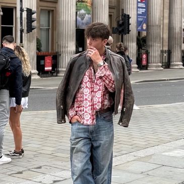 A young man in a patterned shirt and jacket stands on a city sidewalk.