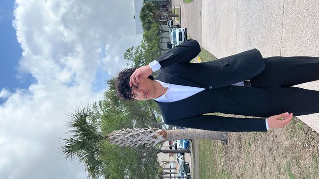 Young man in a black suit standing outdoors by palm trees.