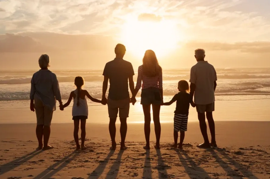 a family of three generations, standing on a beach, looking at the water.