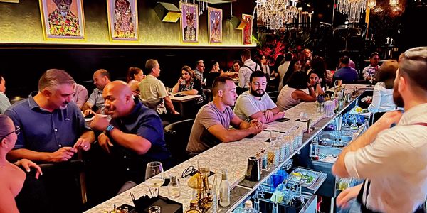 People socializing and enjoying drinks at a stylish, crowded bar with chandeliers.
