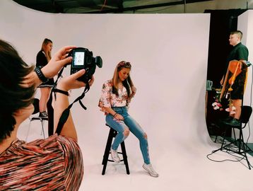 Photographer taking a picture of a girl sat on a chair in a white photography studio. 