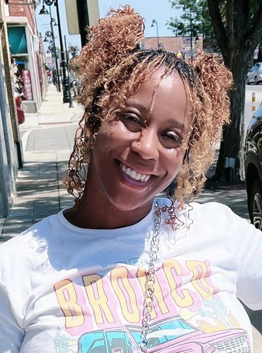 A smiling woman with curly hair styled in two buns outdoors on a sunny day.