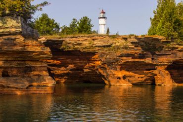 White lighthouse atop rocky cliffs with water below and trees in the background.