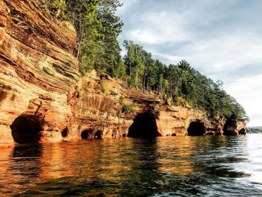 Rocky cliffs with caves reflect in calm water under a cloudy sky.