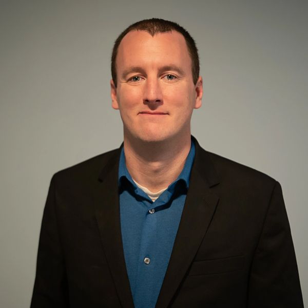 Man in a black blazer and blue shirt smiling slightly.