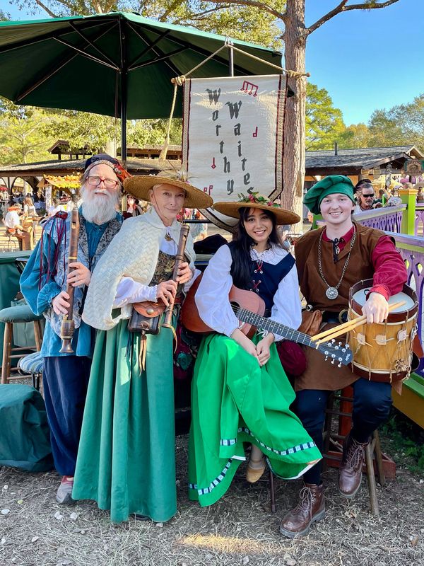 Four musicians in medieval attire at a fair, holding instruments and smiling.