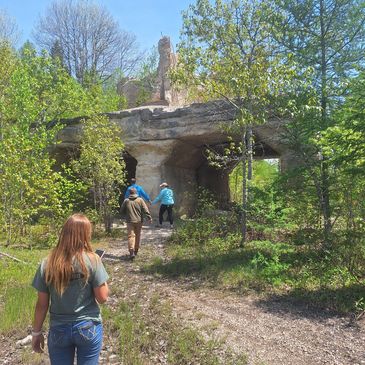 People hiking toward a stone cave in a forest.