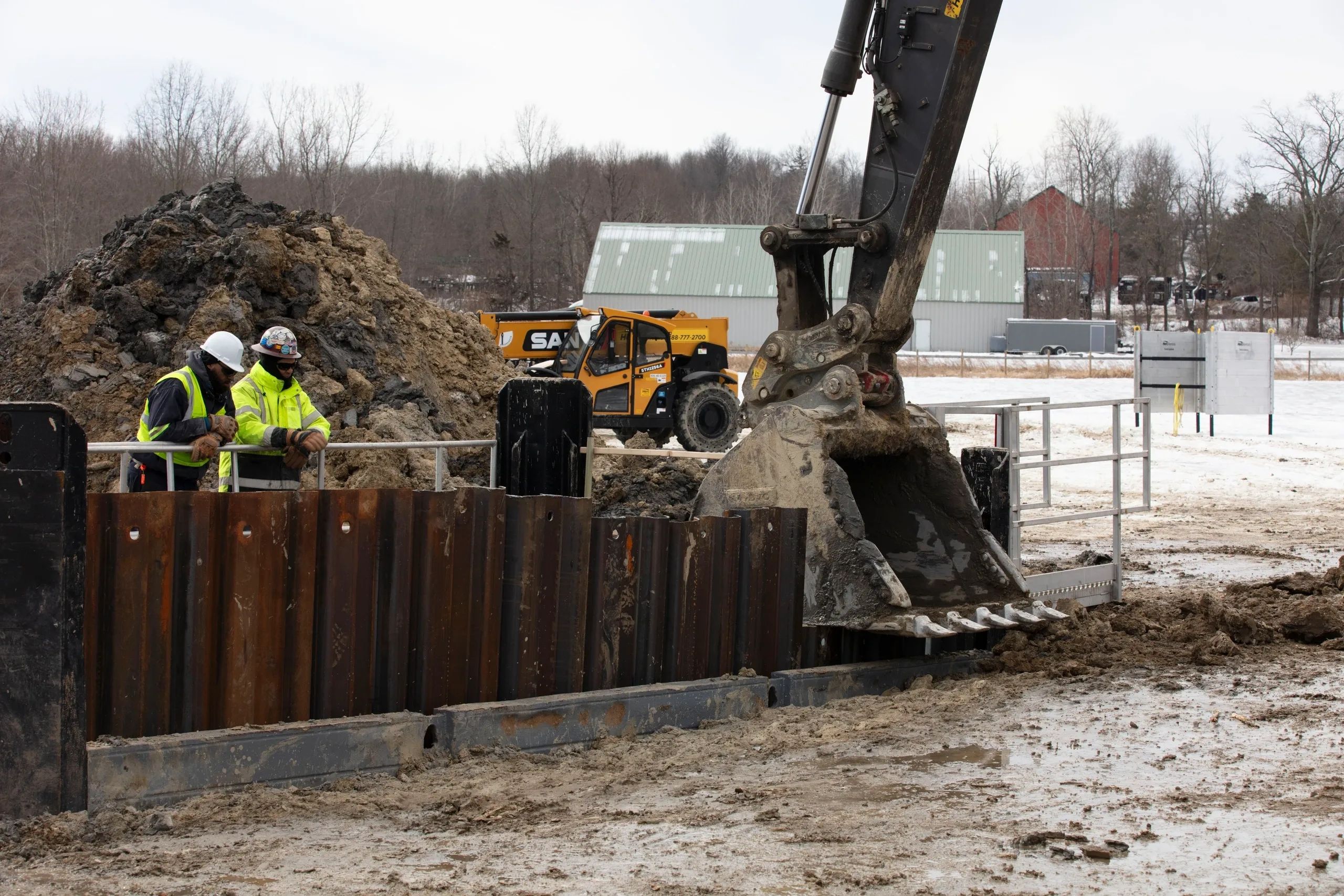 Two construction workers observe an excavator at a muddy outdoor site.