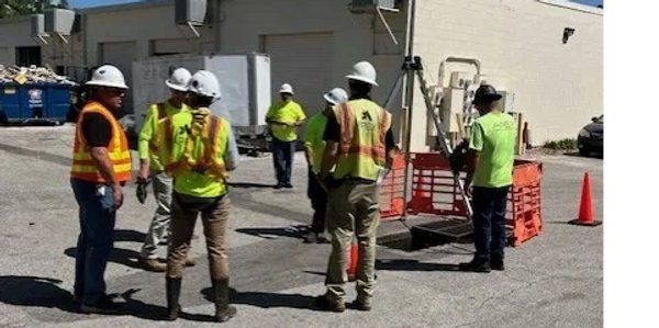 Construction workers in safety gear gathered near an open manhole on a sunny day.