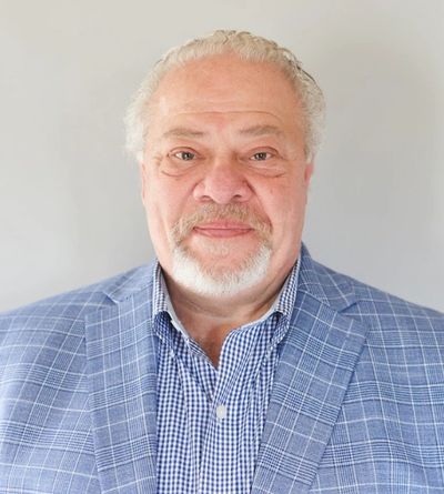 Portrait of an older man in a blue checkered blazer and shirt against a neutral background.