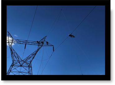Workers performing maintenance on a high-voltage power line tower against a clear blue sky.