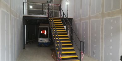 A small mezzanine floor and straight steel staircase with yellow tread nosings and handrail. 