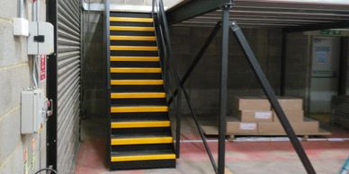 Black straight single flight steel staircase with bright yellow nosings and adjacent mezzanine.