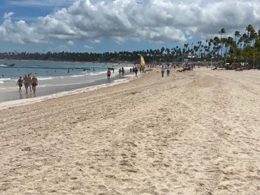Crowded beach with people walking and swimming under partly cloudy sky.