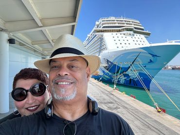 Couple smiling in front of a docked cruise ship on a sunny day.