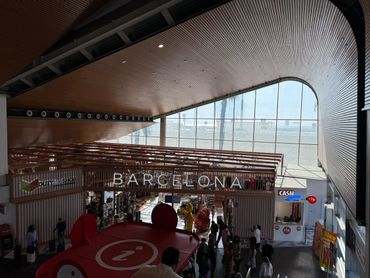 Interior view of Barcelona airport terminal with duty-free shops and people.