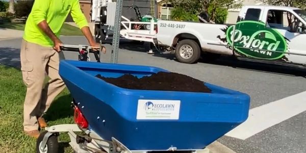 Man uses a blue spreader to distribute soil on a lawn.