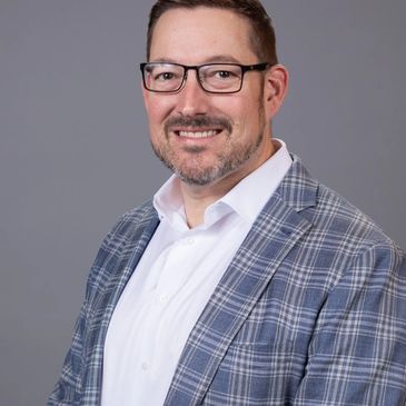 Smiling man in glasses and plaid blazer against a gray background.