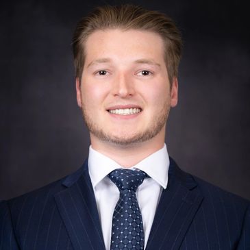 Professional headshot of a young man in a suit and tie against a dark background.