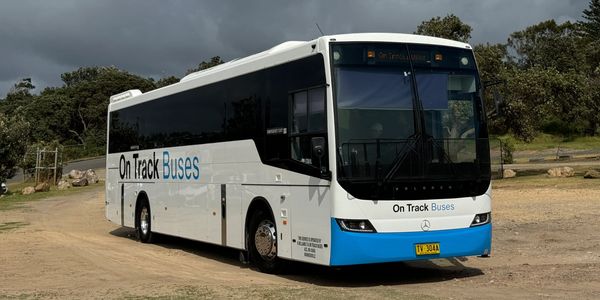 A white and blue On Track Buses coach bus parked on a dirt road under a cloudy sky.