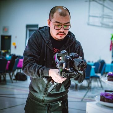 A photographer films a purple-themed cake and cupcakes at an indoor event.